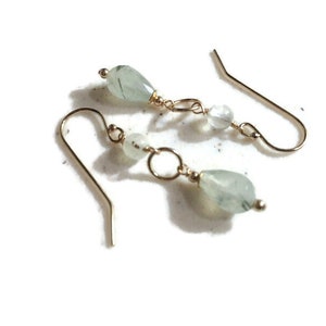 May include: A pair of gold earrings with green and white gemstone beads. The earrings are made with a simple design and feature a small, round bead and a larger, teardrop-shaped bead.
