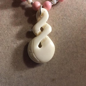 May include: A white carved pendant with a knot design hangs from a pink and white beaded necklace.