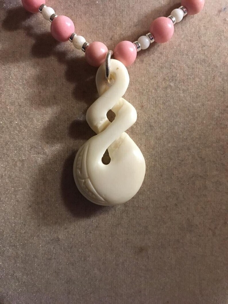 May include: A white carved pendant with a knot design hangs from a pink and white beaded necklace.