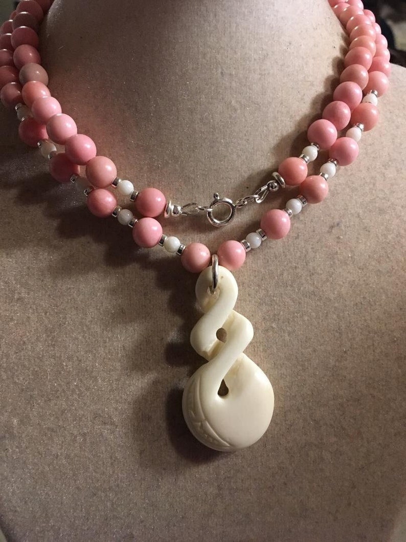 May include: A double strand necklace with pink and white beads and a carved bone pendant. The pendant is a twisted design with a hole in the center.