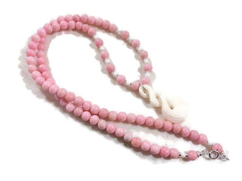 May include: A pink and white beaded necklace with a carved white bone pendant. The pendant is shaped like a knot or a swirl.