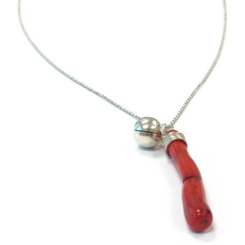 Op de afbeelding: Een zilveren ketting met een klein zilveren belletje en een lange, rode koraal hanger.