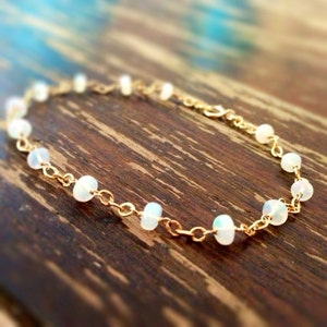 May include: A delicate gold chain bracelet with small white opal beads. The bracelet is on a wooden surface.