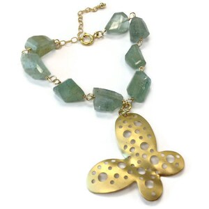 Puede incluir: Un collar de oro con un gran colgante de mariposa recortada. El collar est&aacute; hecho con una cadena y presenta cuentas de piedras preciosas de color verde claro.