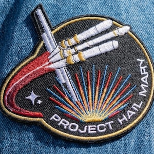 May include: Embroidered patch with a space-themed design. The patch features a black background with a rocket and the text "PROJECT HAIL MARY". The design includes a red and white trail, a star, and the words "HAIL MARY" in white.