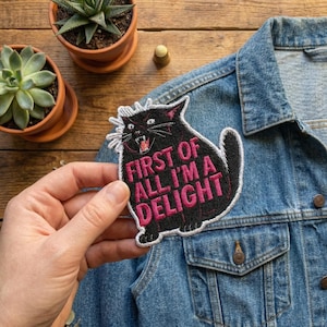 May include: Black cat patch with the text "FIRST OF ALL I'M A DELIGHT" in pink. The patch is being held over a denim jacket. The cat has white whiskers and a white outline.
