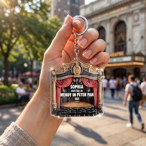 May include: A clear acrylic keychain featuring a theater stage design. The stage has red curtains, a wooden floor, and black seating. Text on the keychain reads "SOPHIA starring as WENDY IN PETER PAN 2025". The keychain is attached to a silver keyring.