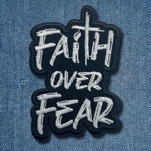 May include: Embroidered patch with the words "Faith Over Fear" in white script on a black background. The patch is in a unique shape and is set against a blue denim background.