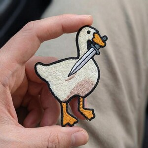 May include: Embroidered patch featuring a white goose with a black outline, holding a silver dagger in its beak. The goose has orange feet and a light brown chest. The patch is held in a hand.