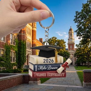 May include: A graduation-themed keychain featuring a black mortarboard hat with a gold tassel atop stacked books. The books display the year "2026", the name "Mike Brown", and "Purdue University". A rolled diploma with a red ribbon is also included.