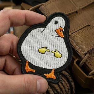 May include: Embroidered patch featuring a cartoon duck with a white body, black outline, and orange beak and feet. The duck is gesturing with yellow hands. The patch is held against a brown background.