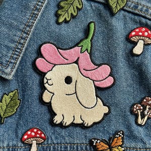 May include: Embroidered patch featuring a cream-colored bunny with a pink flower on its head. The patch is surrounded by other embroidered patches of mushrooms, leaves, and a butterfly, all on a blue denim background.