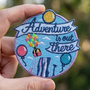 May include: Embroidered patch with the text "Adventure is out there" in a banner. The patch features a colorful hot air balloon, two balloons, and a yellow sun. The patch is round with a light blue background.