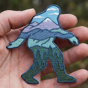 May include: Embroidered patch in the shape of a Bigfoot, featuring a mountain and forest landscape design. The patch is in shades of blue, green, and purple, with white clouds at the top. The patch is held in a hand.