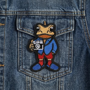 May include: A denim jacket with a pocket featuring an embroidered patch. The patch depicts a cartoon frog wearing a blue jacket, red pants, and blue shoes, holding a camera. The frog has a tan head and a black outline.