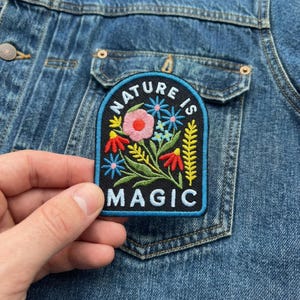 May include: Embroidered patch with the words "NATURE IS MAGIC" and a floral design in red, pink, blue, and yellow. The patch is on a black background with a blue border. The patch is being held up against a denim jacket.