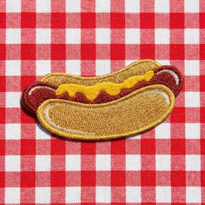 May include: Embroidered patch of a hot dog with mustard on a red and white checkered background. The hot dog features a brown bun, a red hot dog, and yellow mustard. The patch is likely for clothing or accessories.