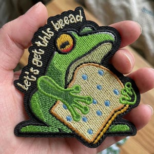 May include: Embroidered patch featuring a green frog holding a piece of bread with blue dots. The patch has the text "let's get this bread" in black lettering. The frog has a yellow eye and is outlined in black.