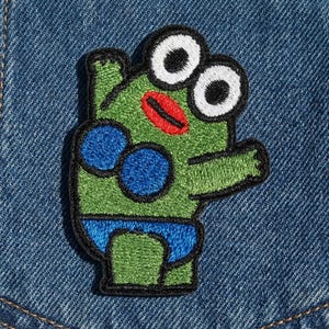 May include: Embroidered patch featuring a cartoon frog with large white eyes, a red mouth, and blue spots. The frog is green with a black outline and wears blue swim trunks. The patch is on a denim background.