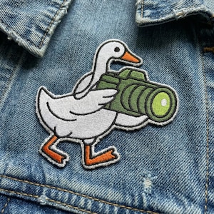May include: Embroidered patch featuring a white goose with an orange beak and feet, holding a green camera. The patch is outlined in black and is affixed to a denim jacket. The goose is walking.