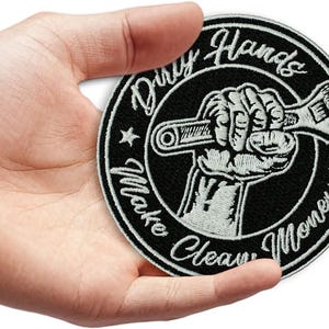 May include: Black and white embroidered patch with the text "Dirty Hands Make Clean Money." The patch features a hand gripping a wrench, surrounded by a circular border with stars. The design is on a black background.