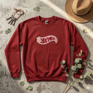 Może przedstawiać: Bordowa bluza z napisem "Hot Mess" w białym i różowym wzorze. Bluza z okrągłym dekoltem jest prezentowana na teksturowanej powierzchni z kluczami, słomkowym kapeluszem i zielenią.