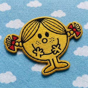 May include: Embroidered yellow patch featuring a cartoon character with black outlines, freckles, and red bows. The character has a surprised expression with open arms and legs. The patch is on a light blue background with white clouds.