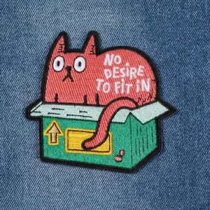 May include: Embroidered patch featuring a red cat in a green box with the text "No Desire To Fit In." The cat has large eyes and a long tail. The patch is on a blue denim background.