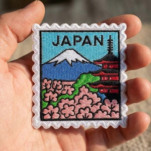 May include: Embroidered patch featuring a design inspired by Japan. The patch depicts Mount Fuji, a pagoda, and cherry blossoms, with the word "JAPAN" at the top. The patch has a white scalloped border.
