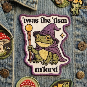 May include: Embroidered patch featuring a green frog wearing a purple wizard hat and cape, holding a staff. The patch has the text "'twas the 'fism m'lord" in black. The patch is on a denim jacket.
