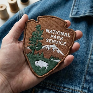 May include: A brown and green embroidered patch with the text "NATIONAL PARK SERVICE". The patch features a tree, mountains, and a bison. The patch is held against a denim jacket.