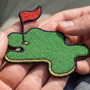 May include: Embroidered patch featuring a green golf course with a red flag. The patch has a black border with yellow accents. The patch is held in a person's hands.