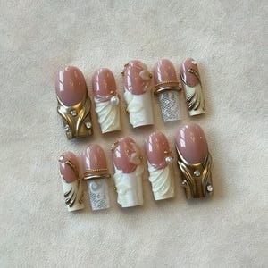 May include: A set of ten artificial nails with a light pink oval shape. Some nails feature gold-colored metal accents with rhinestones, while others have white lace and pearl details. The nails are designed for a glamorous look.