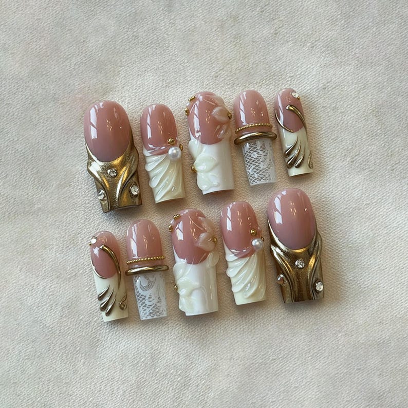 May include: A set of ten artificial nails with a light pink oval shape. Some nails feature gold-colored metal accents with rhinestones, while others have white lace and pearl details. The nails are designed for a glamorous look.
