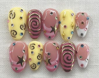 Op de afbeelding: Een set van tien kunstnagels met verschillende ontwerpen. Sommige nagels zijn geel met spiraal- en stipontwerpen, andere roze met gouden ster- en streepaccenten. Verschillende nagels hebben kleurrijke strass-steentjes.