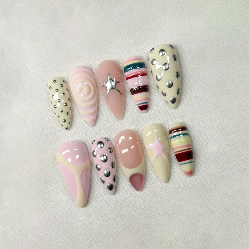 May include: A set of ten long, almond-shaped artificial nails. The nails feature various designs, including swirls, stripes, stars, and silver studs. Colors include pink, cream, and red.