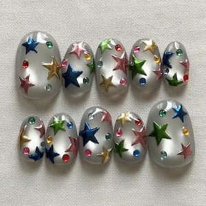 May include: Set of ten silver-colored, oval-shaped artificial nails. Each nail is decorated with colorful star-shaped embellishments and small, round, jewel-like accents in various colors. The stars are in shades of blue, green, pink, and gold.