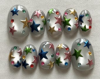 Op de afbeelding: Set van tien zilverkleurige, ovale kunstnagels. Elke nagel is versierd met kleurrijke stervormige versieringen en kleine, ronde, juweelachtige accenten in verschillende kleuren. De sterren zijn in tinten blauw, groen, roze en goud.