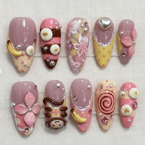 May include: A collection of ten artificial nails with diverse designs. The nails showcase pink, yellow, and brown hues, adorned with pearls, flowers, and a heart. Some nails feature banana and monkey designs, offering a playful aesthetic.