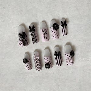 May include: A set of ten artificial nails with various designs. Some nails are pink with black polka dots, others are pink with black stripes, and one is pink with the word "Doll". The nails feature black bows, lace, and buttons.