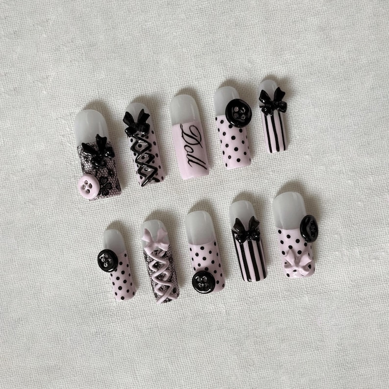 May include: A set of ten artificial nails with various designs. Some nails are pink with black polka dots, others are pink with black stripes, and one is pink with the word "Doll". The nails feature black bows, lace, and buttons.