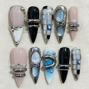 May include: A collection of ten long, pointed artificial nails. The nails feature various designs, including solid colors like black and pink, and intricate patterns with silver accents, glitter, and geometric shapes. Some nails have silver rings.