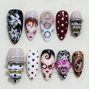 May include: A set of ten artificial nails with diverse designs. The designs include floral patterns, faces, polka dots, and abstract art. Some nails feature embellishments like pearls and metal rings. The colour palette includes black, white, red, and nude tones.