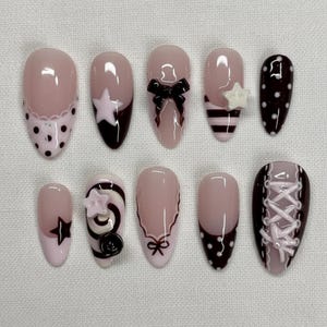 Puede incluir: Un juego de diez uñas postizas en forma de almendra con acabado brillante en tonos rosa pálido, negro y blanco. Los diseños incluyen lunares, rayas, un lazo negro, una estrella blanca, una espiral, un detalle de cordones tipo corsé y bordes festoneados.