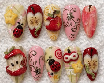 May include: A set of ten almond-shaped press-on nails featuring 3D designs including red apples, ladybugs, yellow stars, and swirling patterns. The nails display various finishes such as plaid backgrounds, metallic gold accents, and glossy red fruit.