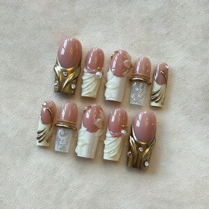 May include: A set of ten artificial nails with a light pink base and various decorative elements. Some nails feature gold-colored accents, rhinestones, and white floral or lace designs. The nails are arranged on a light-colored surface.