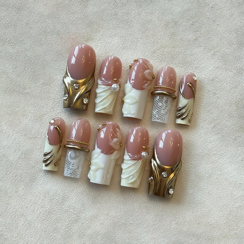 May include: A set of ten artificial nails with a light pink base and various decorative elements. Some nails feature gold-colored accents, rhinestones, and white floral or lace designs. The nails are arranged on a light-colored surface.