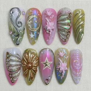 May include: A set of ten almond-shaped press-on nails featuring intricate 3D designs including metallic silver swirls, a crescent moon with stars, pink floral reliefs, iridescent butterfly wings, gold star accents, and swirling textures in pastel tones.