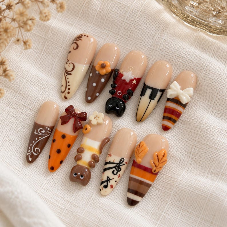 May include: A set of ten almond-shaped press-on nails featuring various designs including brown swirls, polka dots, a ladybug, piano keys, a white bow, orange stripes, a small cat figure, musical notes, and orange autumn leaves.