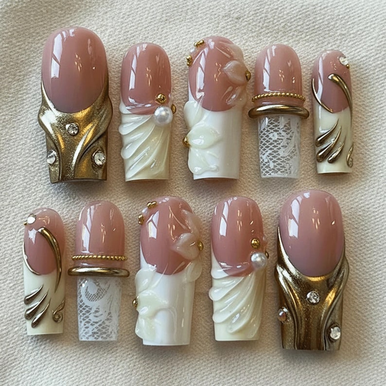 May include: A collection of ten artificial nails with various designs. The nails feature a glossy, light pink color with gold, white, and pearl accents. Some nails have gold embellishments, while others have lace or floral details.